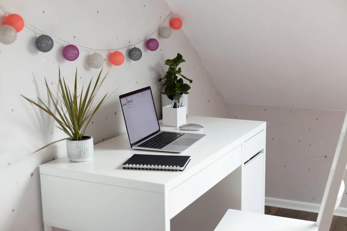 A compact white desk with a laptop, notebook and potted plants beneath a colorful globe garland.
