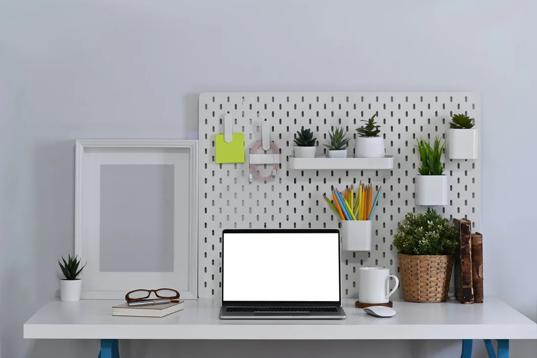 A white desk with a laptop, pencil cup and small plants below a white pegboard.