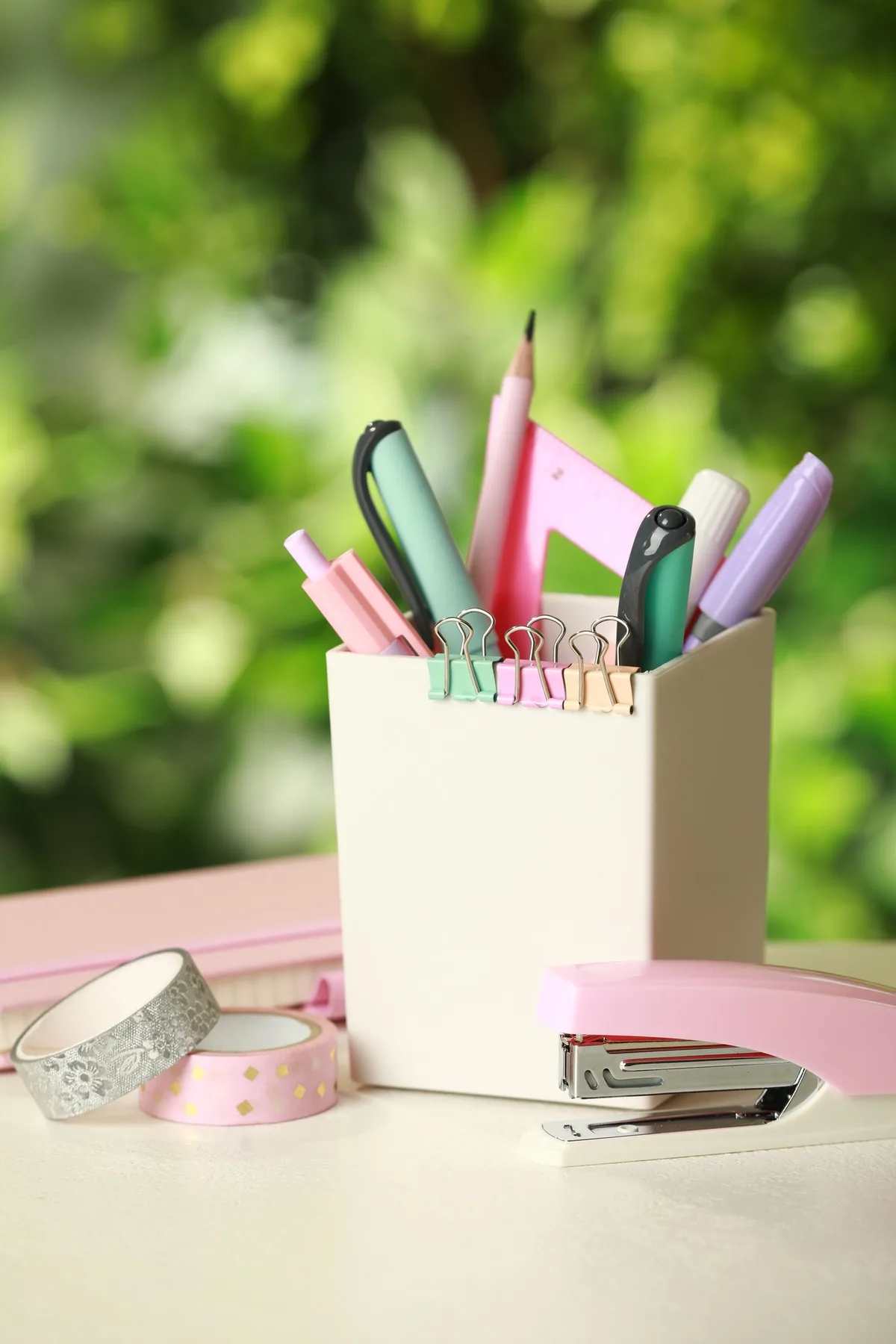 A white pen holder with pastel pens, pencils and clips beside a pink stapler and decorative tape.