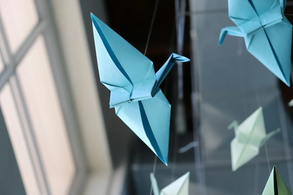Blue and pale origami cranes suspended from fine threads near a window.