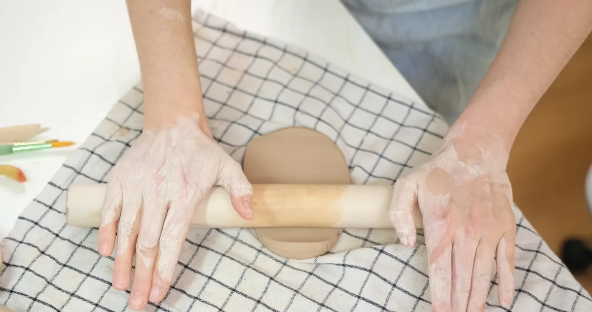Hands rolling a supported slab of clay on a checked cloth.