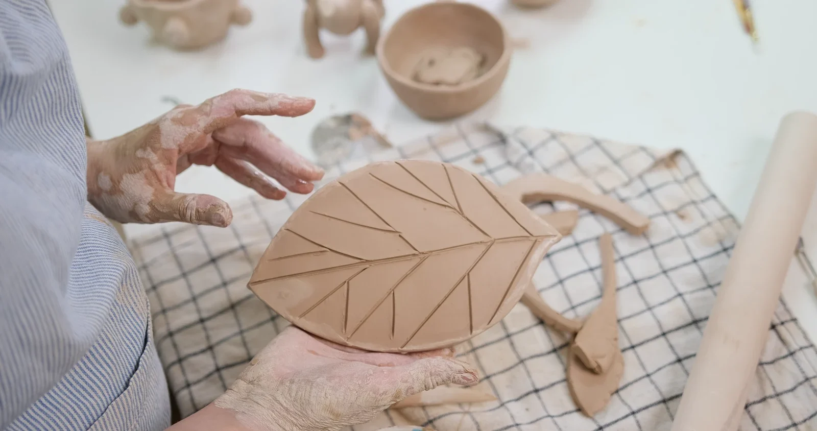 Hands supporting a broad clay leaf with a center vein and diagonal branching grooves.