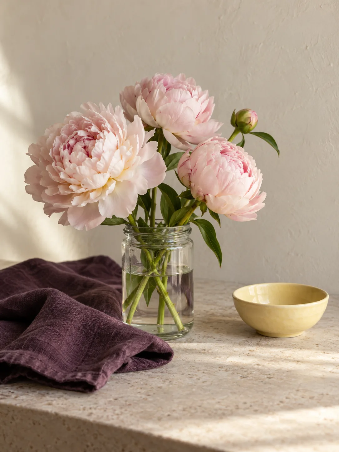Blush peonies in a clear jar beside deep plum linen and a small butter-yellow ceramic bowl on pale stone.